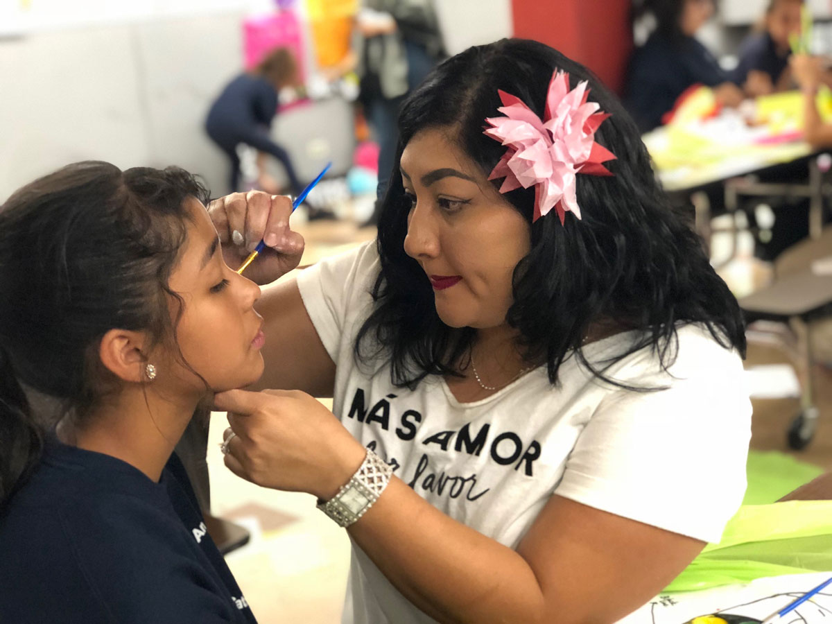 A madrina at Annunciation School in Denver, CO, painting the face of a student for the Dia de los Muertos celebration that madrinas team organized.