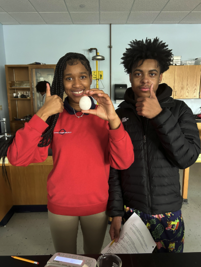 Two students (female in red sweatshirt on left and male in black puffer jacket on right) posing for a photo with the female student holding an egg. Both students are giving a thumbs up to the camera