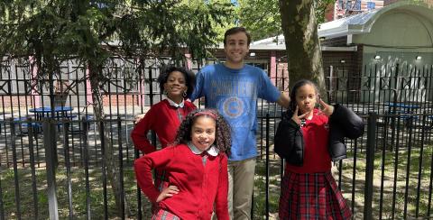 Image of Jack Hutter in blue tshirt posing with three of his students in red sweaters and red plaid skirts: two girls to his left and one to his right.