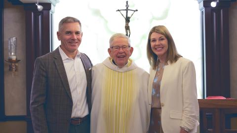 Fr. Joe Carey, CSC in white clerical robes stands between a smiling man in a dark grey suit and a smiling woman in a white jacket (right) 