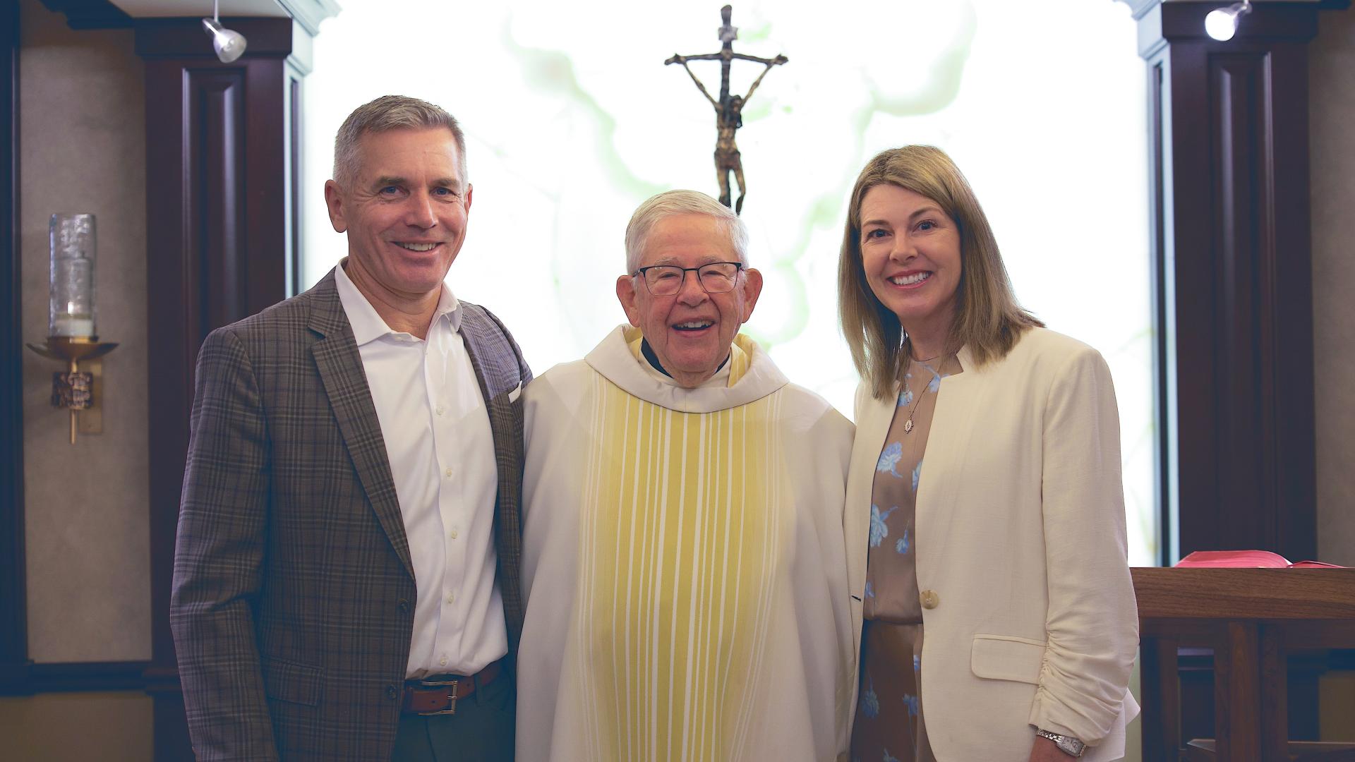 Fr. Joe Carey, CSC,  in white clerical robes stands between a smiling man in a dark grey suit and a smiling woman in a white jacket (right) 
